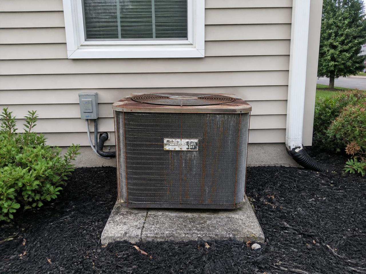 Same-location before photo: corroded outdoor AC condenser against the home's beige siding, mulch bed, and downspout before replacement