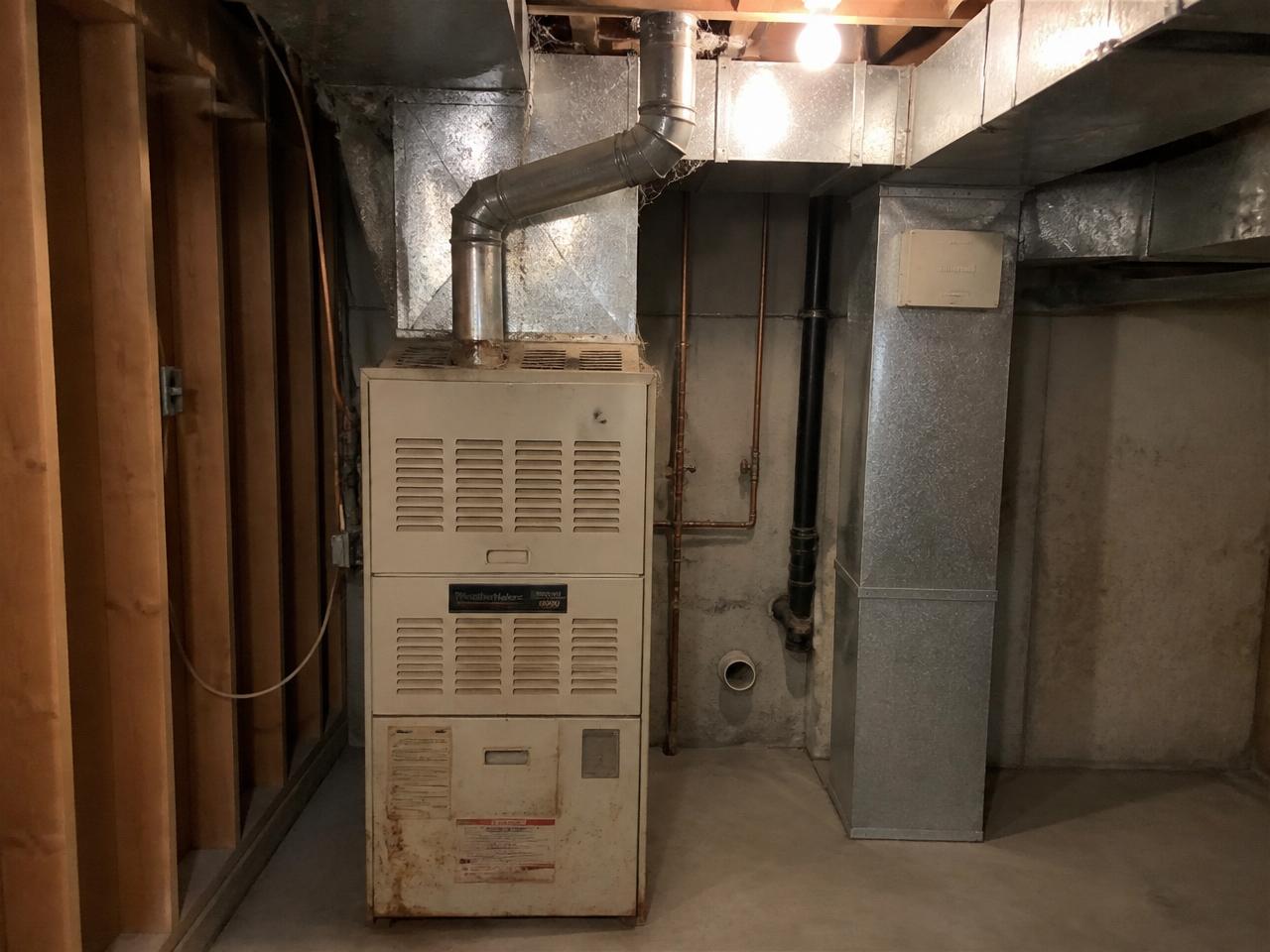 Same-location before photo: old dusty gas furnace in the basement utility corner with the same studs, ducts, copper lines, and PVC vent